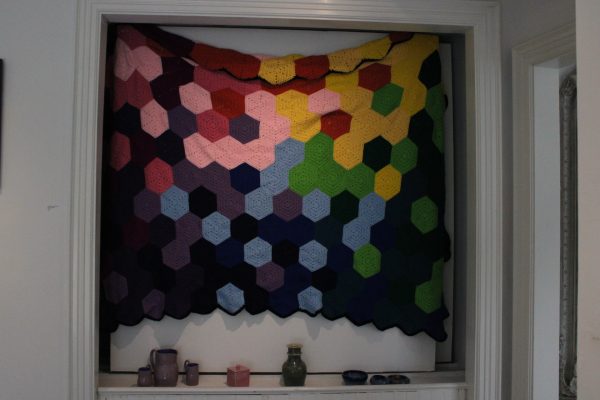 Kassidy Klopotoski crocheted blanket hangs on display above pottery on display.